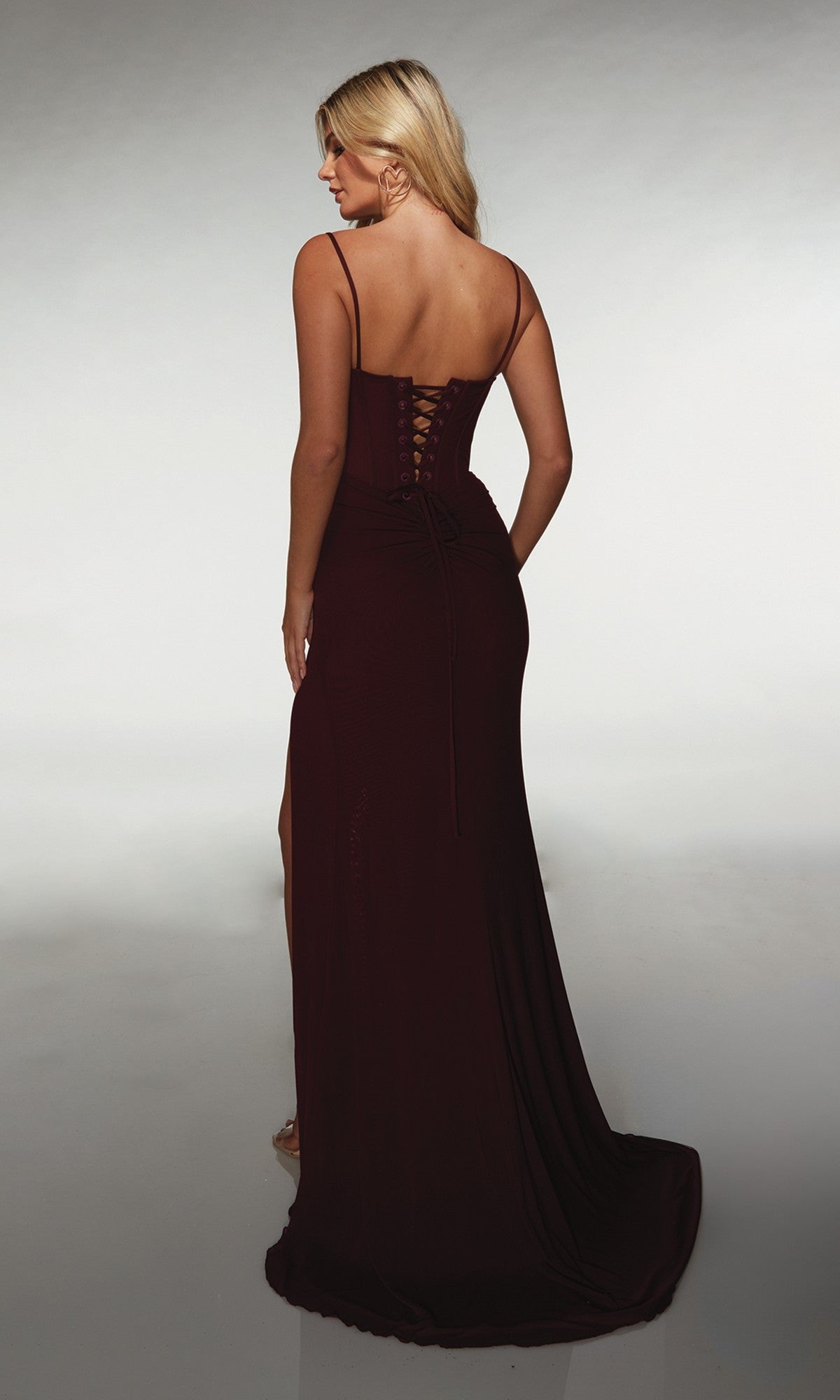 Woman in the Alyce 62002, a floor-length dark burgundy prom dress featuring spaghetti straps, a lace-up corset back, and a side slit, is standing and facing away from the camera.